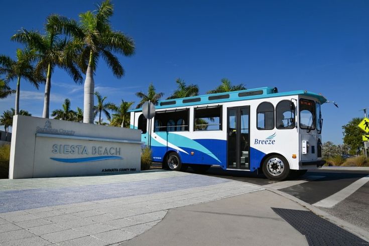 Siesta Key Breeze Trolley parked near the Siesta Beach entrance on a sunny day, with palm trees and a clear blue sky in the background.