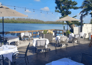 Outdoor waterfront dining area with white tablecloth-covered tables, patio umbrellas, and string lights overlooking a calm bay on a sunny day in Siesta Key.