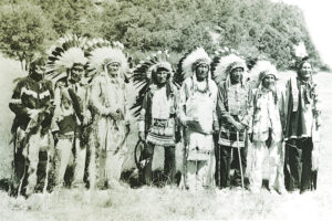 A black-and-white photograph of eight Native American men wearing feathered headdresses and traditional clothing, standing in a row outdoors.