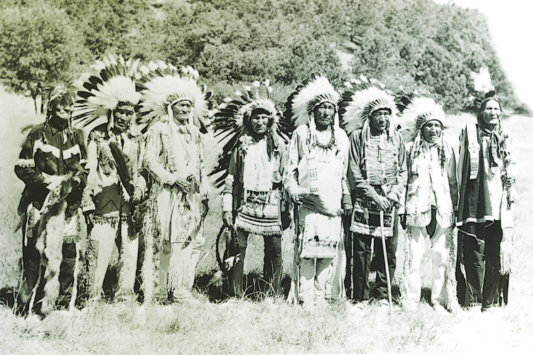 A black-and-white photograph of eight Native American men wearing feathered headdresses and traditional clothing, standing in a row outdoors.