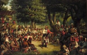 A large gathering of Native American men and women in traditional dress listen to a leader speaking in an outdoor council beneath tall trees.