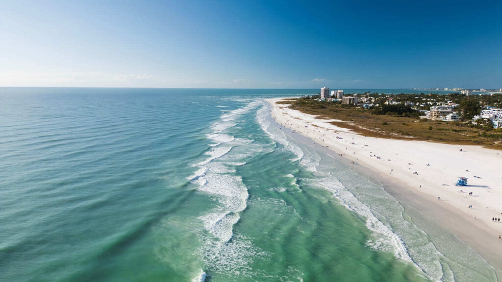Aerial view of Siesta Key Beach with turquoise waves rolling onto the wide white sand shoreline, and buildings along the coast under a clear blue sky.