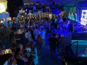 Indoor view of a Halloween party at The Beach Club Siesta Key, showing a live band performing for a large, dancing crowd under vibrant blue lights.