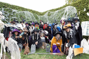 Joyful group of costumed witches celebrating Halloween amidst a spooky outdoor display with white spiderwebs and tombstones.