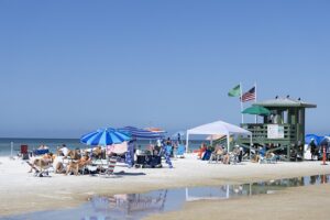 Beachgoers relax under colorful umbrellas near a green lifeguard tower on Siesta Beach in Sarasota, Florida, on a bright sunny day.