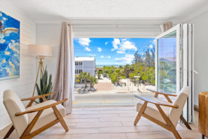 Bright coastal vacation rental sitting area with two modern armchairs and large open window overlooking a palm-lined street and partial Gulf view in Siesta Key, Florida.