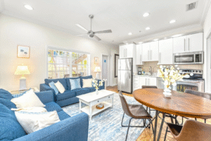 Bright, open-concept vacation rental living room and kitchen in Siesta Key featuring blue sofas, white cabinetry, stainless steel appliances, round wooden dining table, and large windows with natural light.