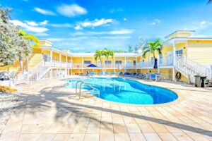 Resort-style vacation rental property in Siesta Key featuring a large outdoor swimming pool, yellow coastal-style buildings, white railings, palm trees, and poolside lounge chairs under a sunny blue sky.
