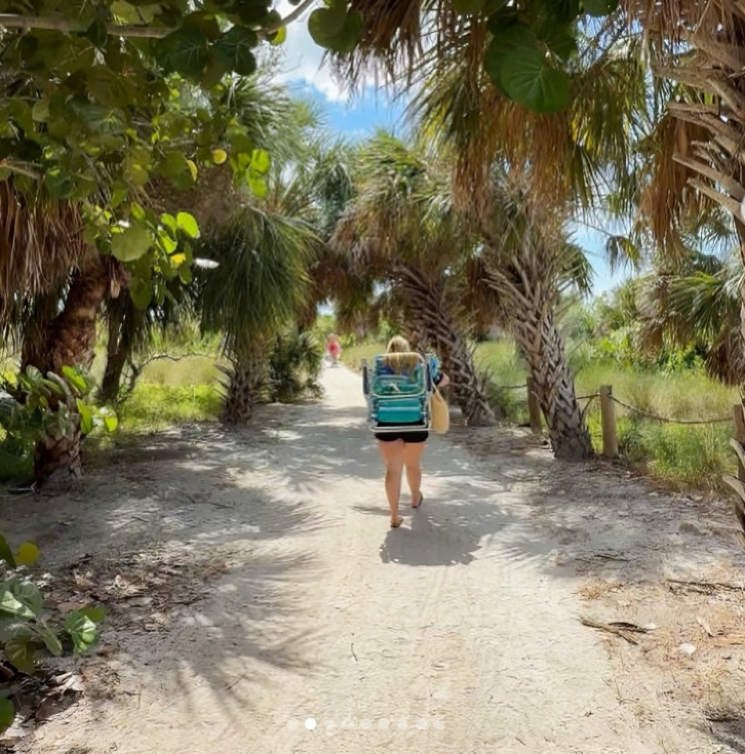 A sandy path lined with palm trees and lush greenery leads toward the beach, where a person carrying beach gear walks under the shade on a sunny day.