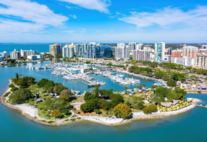 Aerial view of Bayfront Park in Sarasota, Florida, featuring waterfront walking paths, a marina filled with boats, Sarasota Bay, and the downtown skyline.