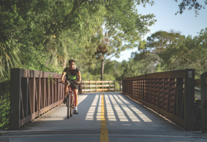 Cyclist riding across a bridge along The Legacy Trail surrounded by lush greenery in Sarasota, Florida.
